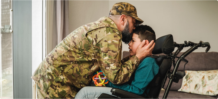 A military service member in uniform kisses a child who uses a wheelchair on the forehead.