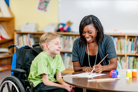 A child who uses a wheelchair paints and laughs with a special education professional.