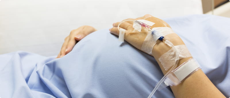 A pregnant woman in a medical gown lying down with an IV line in her hand.