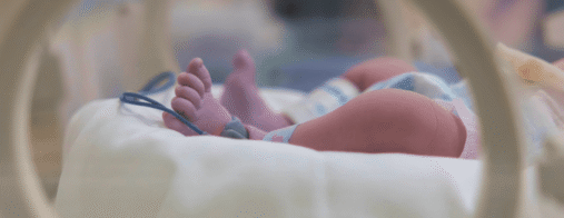 A newborn lies in a hospital bassinet with a monitoring band attached to the foot.