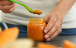 A person scoops baby food from a glass jar with a plastic spoon.