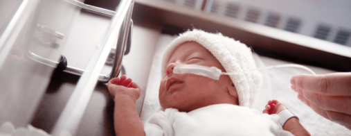 A newborn wearing a knit cap lies in a hospital bassinet with nasal tubing for oxygen therapy.