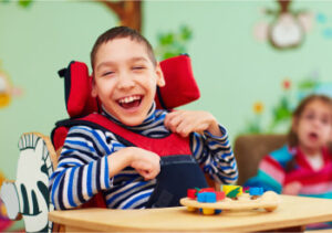 A child with cerebral palsy plays and laughs at a desk in a classroom.