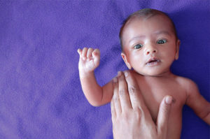 An adult hand touches a baby's right shoulder.