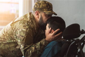 A man in military fatigues kisses a boy in a wheelchair on his head 