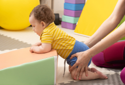 A baby in therapy is supported by an adult while practicing crawling on soft play equipment.