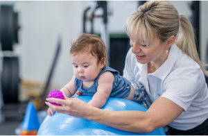 A therapist supporting a baby on a therapy ball while encouraging them to reach for a textured toy.