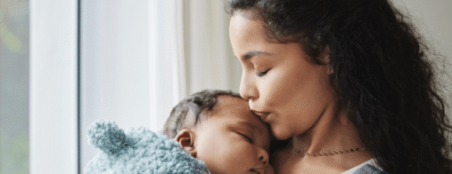 A mother kisses her sleeping baby on the forehead while holding them close.