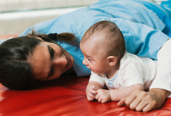 A therapist lies on a padded mat face-to-face with a baby, engaging in a developmental exercise.
