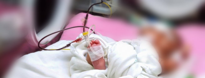 Close-up of a premature baby’s foot with medical tubing and monitoring lines in a neonatal intensive care unit.