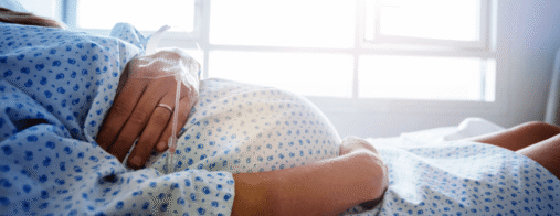 A pregnant woman lies in a hospital bed with one hand on her belly and an IV in the other hand.