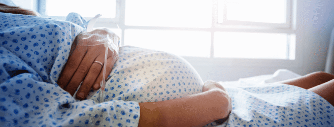 A pregnant woman lies in a hospital bed with one hand on her belly and an IV in the other hand.