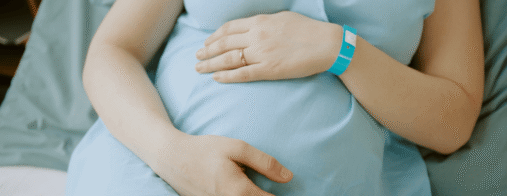 A pregnant woman in a hospital gown rests both hands on her belly while sitting in a hospital bed.
