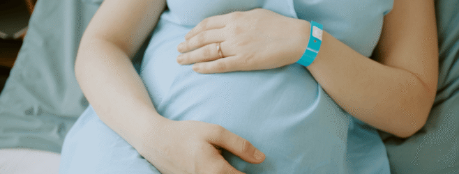 A pregnant woman in a hospital gown rests both hands on her belly while sitting in a hospital bed.