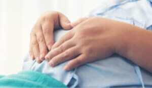 A pregnant mother in a hospital gown holds her hands over the baby she is carrying.