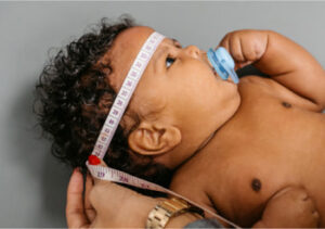 A baby's head is measured just above the eyebrows and ears with a measuring tape.