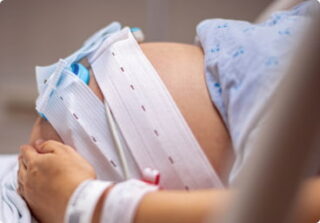 A pregnant woman lies on a hospital bed with a fetal monitoring belt wrapped around her belly.