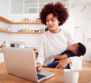 A mother works on a laptop in her kitchen while holding her sleeping baby.