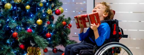 A young boy in a wheelchair shakes a wrapped gift in front of a decorated tree