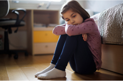A young girl sits on the floor by her bed with her knees pulled close, looking sad and thoughtful.